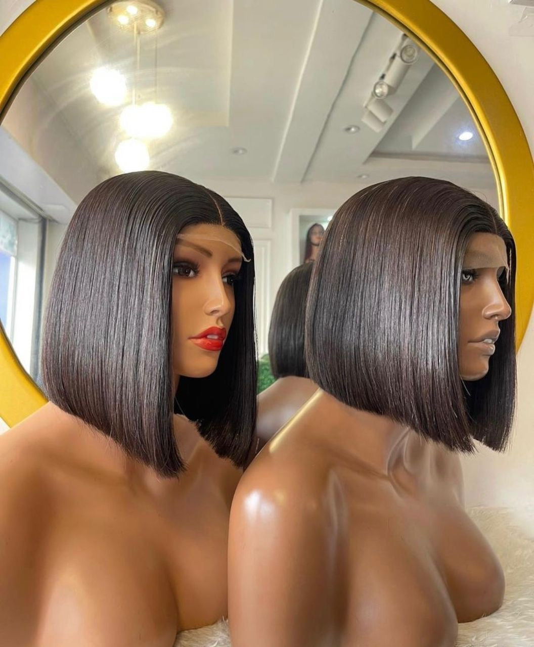 Two mannequins wearing black centre-parted blunt bob wigs in front of a round mirror.